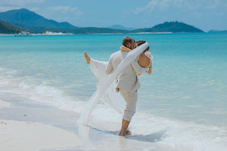 Couple in wedding attire embracing near the shoreline of Whitehaven Beach in the Whitsunday Islands